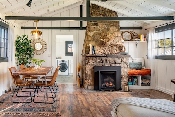 A cozy living room with a stone fireplace, a dining area with a wooden table and chairs, and a view of the kitchen in the background.