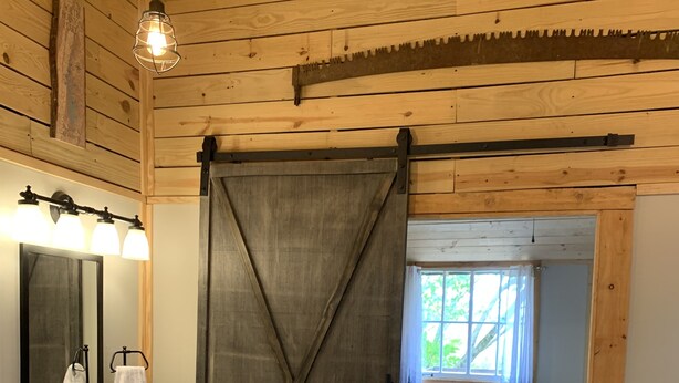Interior de una cabaña de madera con un baño que incluye un lavabo, espejo y toallero. Una puerta corrediza tipo granero da paso a un dormitorio con cama y ventana.