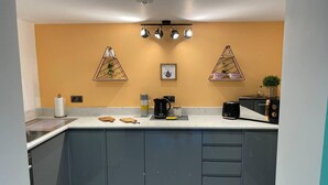 A modern kitchen with grey cabinets, a white countertop, and a yellow accent wall. There are two pendant lights, a coffee maker, and a toaster on the counter. - Hideaway 1-bed Apartment in Herne Bay (Herne Bay)