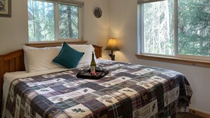 Ein ordentlich bezogenes Bett mit einer Steudecke, eine Nachttischlampe und ein Fenster mit Blick auf einen Wald.
