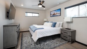 A bedroom with a bed, a nightstand, a TV, a ceiling fan, a window, and a painting on the wall.