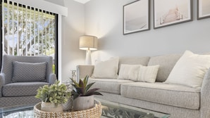 A living room with a grey sofa, a patterned armchair, a glass coffee table, and a lamp. There are framed pictures on the wall and a window with blinds.
