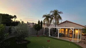 A backyard with a putting green, a covered seating area, and a house in the background.