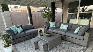 A covered outdoor seating area with a grey sectional sofa, matching ottoman, and patterned rug.