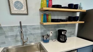 A kitchen with a stainless steel sink, a coffee maker, and a set of colorful glass tumblers on shelves. - WATERFRONT Burnett House Up (Port O'Connor)