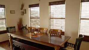 A dining room with a large wooden table, six chairs, and three windows with blinds.
