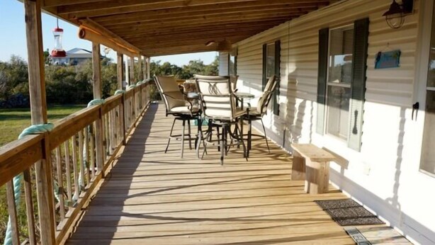 Een houten terras met tuinmeubilair, een welkomstmat en een huis met wit gevelbekleding en luiken.