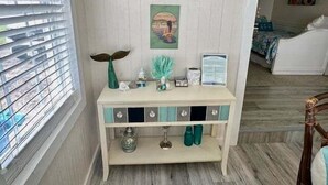 A room with a white wall, a window with blinds, a decorative sign reading "Margaritaville", a small table with teal drawers, and a bed with a white headboard. - SIREN COVE is Close to Beach in POC (Port O'Connor)