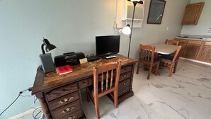 Una scrivania in legno con un computer, una lampada e un cartello con scritto "Thank you for not smoking". Sullo sfondo, una sedia, una TV e un angolo cottura.