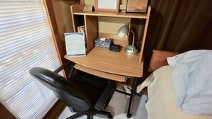 A compact bedroom with a wooden desk, a black office chair, a bed with white bedding, and a window with blinds.