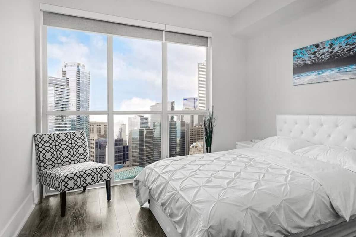 A bedroom with a large window, a white bed, and a chair.