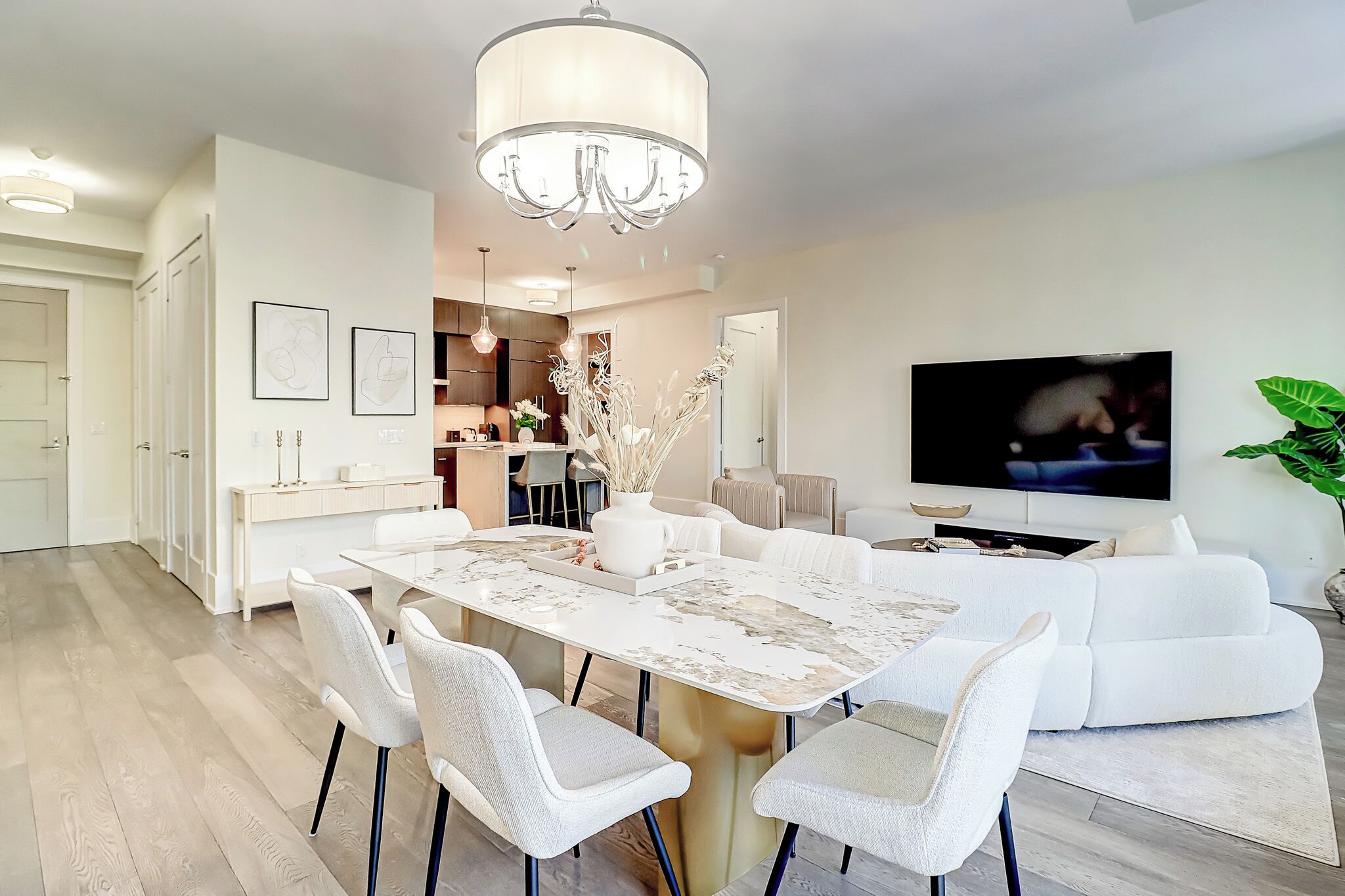 A modern living room with a dining area, a chandelier, a flat-screen TV, and a large plant.