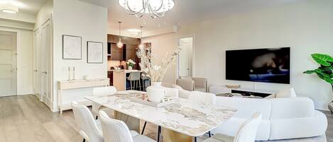 A modern living room with a dining area, a chandelier, a flat-screen TV, and a large plant.