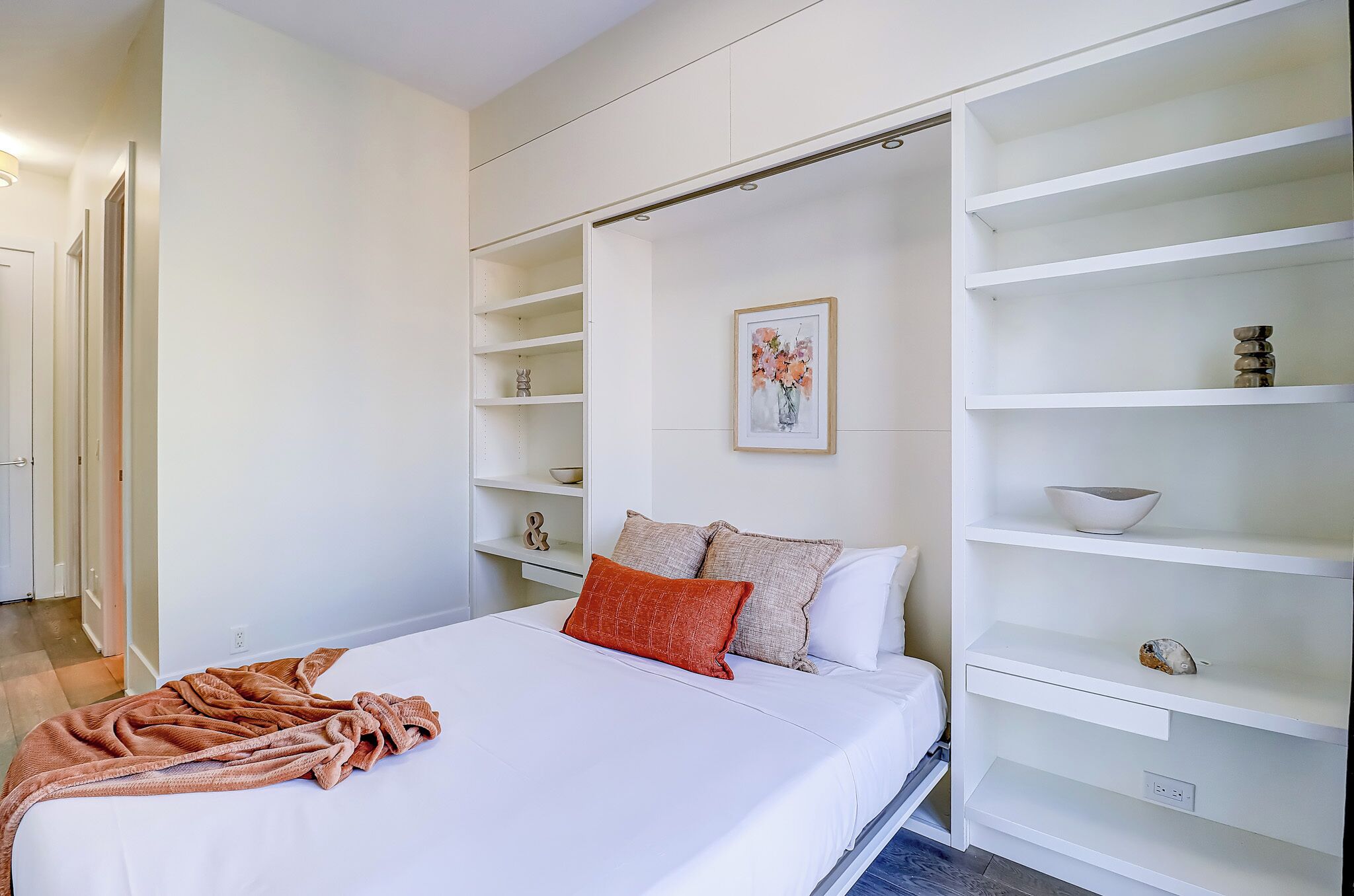 Quarto com cama, paredes brancas, um quadro e estantes embutidas.