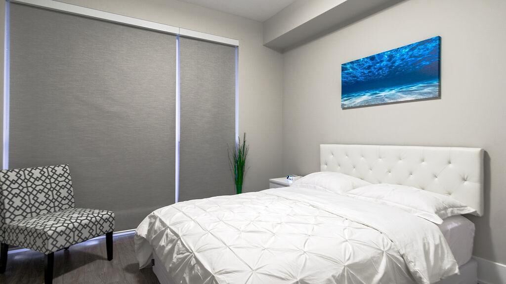 A bedroom with a bed, a chair, a nightstand, a painting, and a window with blinds.