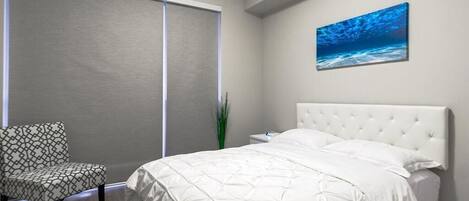 A bedroom with a bed, a chair, a nightstand, a painting, and a window with blinds.