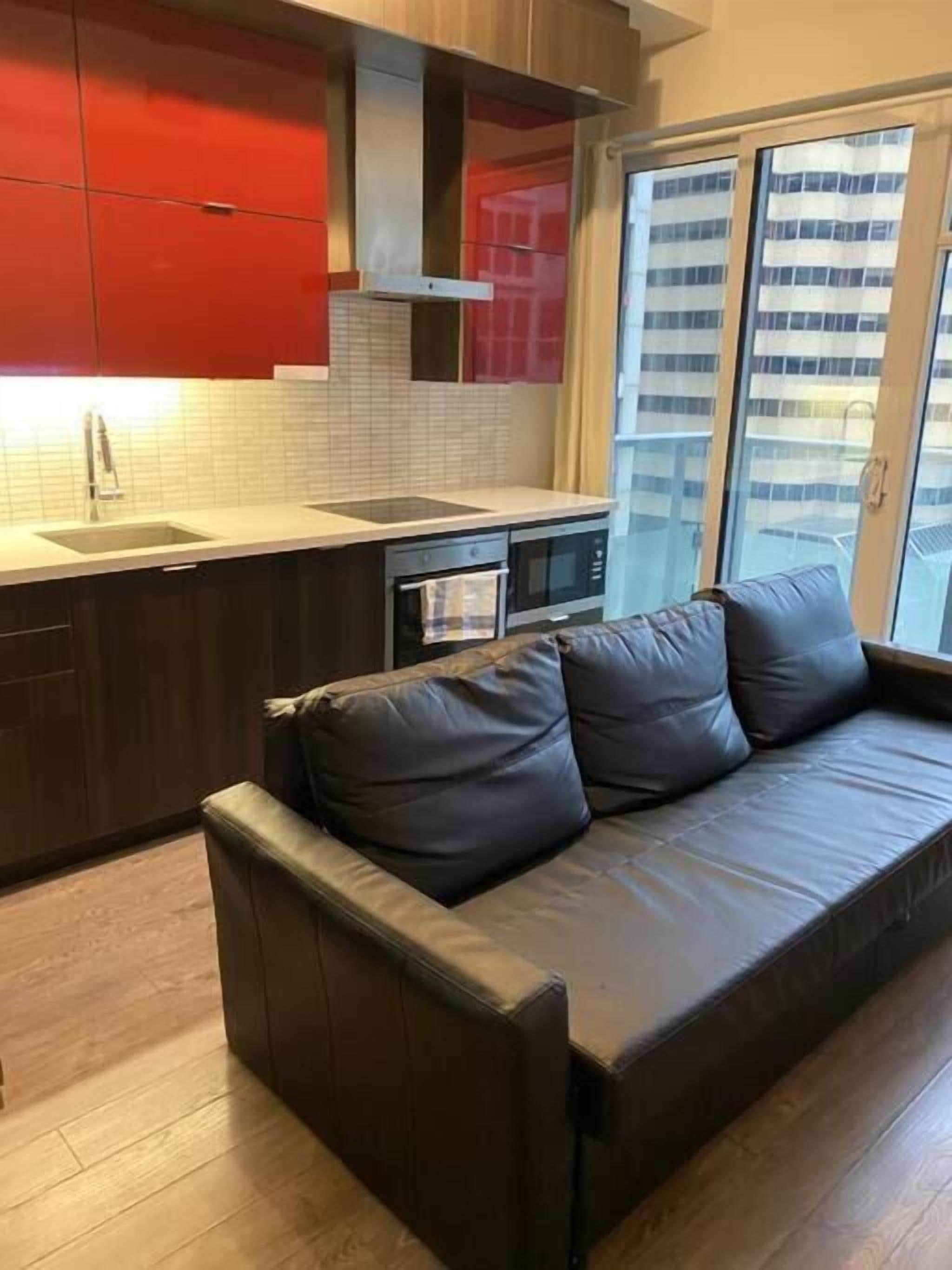 A modern kitchen with red cabinets, a black countertop, and a built-in oven. A brown leather sofa is positioned in front of a large window with a city view.