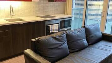 A modern kitchen with red cabinets, a black countertop, and a built-in oven. A brown leather sofa is positioned in front of a large window with a city view.