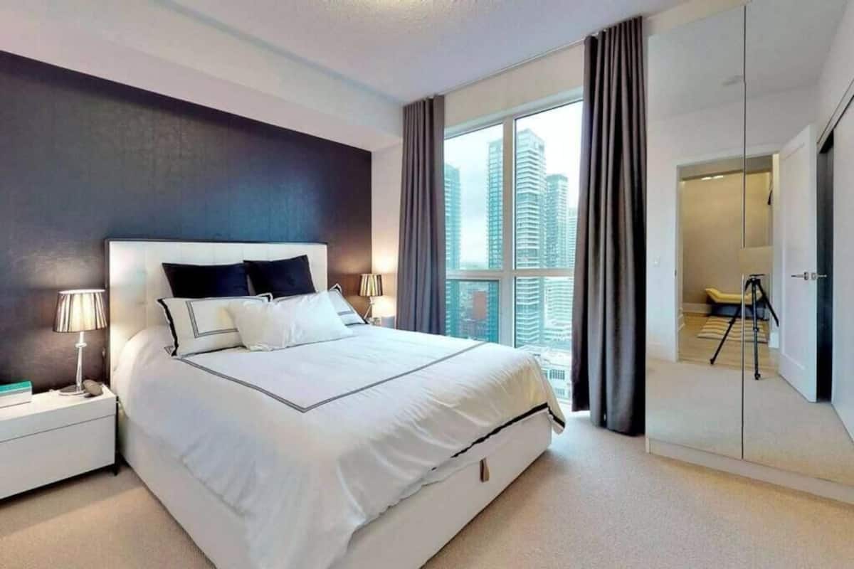 A modern hotel bedroom with a large bed, a nightstand with a lamp, and a view of tall buildings through the window.