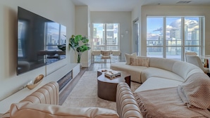 A modern living room with a large flat-screen TV, a beige sofa, a coffee table, and a large window with a city view. - Skyline Suites - Next to MTCC, ACC & CN Tower (Toronto)