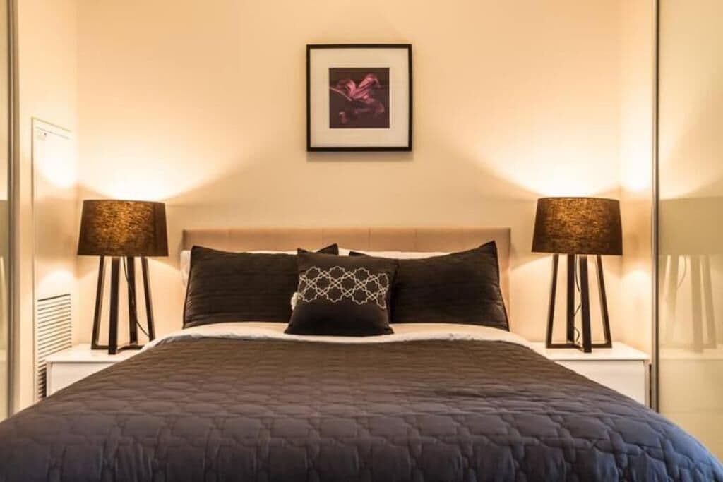 Ein ordentlich bezogenes Bett mit dunkler Decke, zwei Nachttischlampen und einem gerahmten Bild über dem Kopfteil.