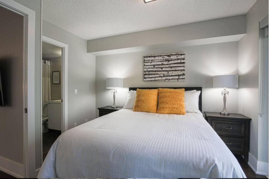A bedroom with a large bed, two bedside tables with lamps, a wall-mounted artwork, and a door leading to a bathroom.