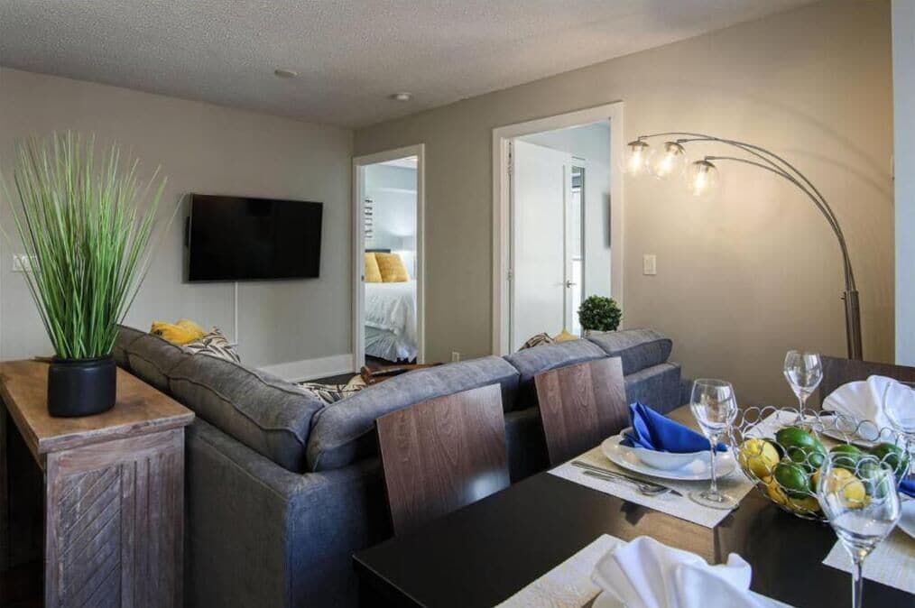 A modern living room with a grey sofa, a wooden side table with a potted plant, a flat-screen TV mounted on the wall, and a dining area with a table set for two.