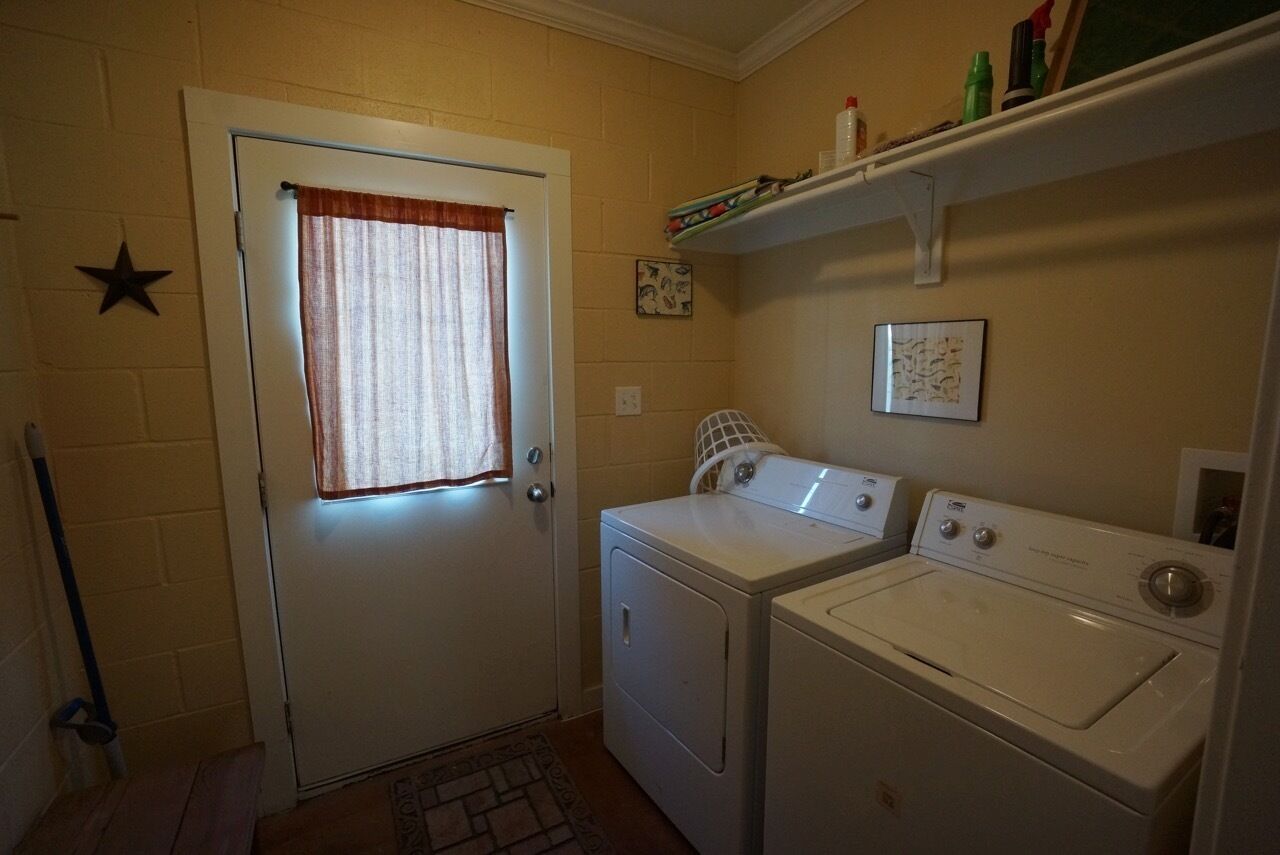 A laundry room with two washing machines, a door with a star decoration, a window with a curtain, and a shelf with various items.