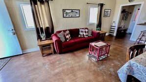 A living room with a red sofa, a wooden coffee table, and a brick wall. There are two windows with curtains, a clock on the wall, and a door leading to another room. - UNCLE WOODY'S #1 in POC-Pets ok but add Fees (Port O'Connor)