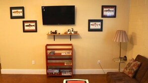Un soggiorno con un divano marrone, una televisione montata a parete, un tavolino con una lampada e una libreria con libri e oggetti decorativi.