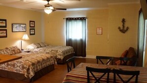 A room with two beds, a dining table with chairs, a ceiling fan, and framed pictures on the walls. - UNCLE WOODY'S #3 in POC-Pets ok but add Fees (Port O'Connor)