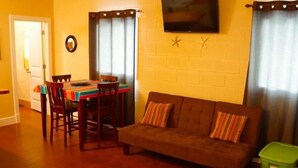 A living room with a sofa, a dining table with chairs, a television mounted on the wall, and a green trash bin. - UNCLE WOODY'S #4 in POC-Pets ok but add Fees (Port O'Connor)