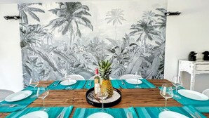Une table à manger dressée avec de l’argenterie, des couverts et des verres, un ananas en guise de centre de table et une bouteille de vin, le tout sur fond de fresque tropicale.