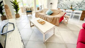 Un salon avec un canapé beige, une table basse blanche, des paniers en osier et une fresque murale aux motifs tropicaux.