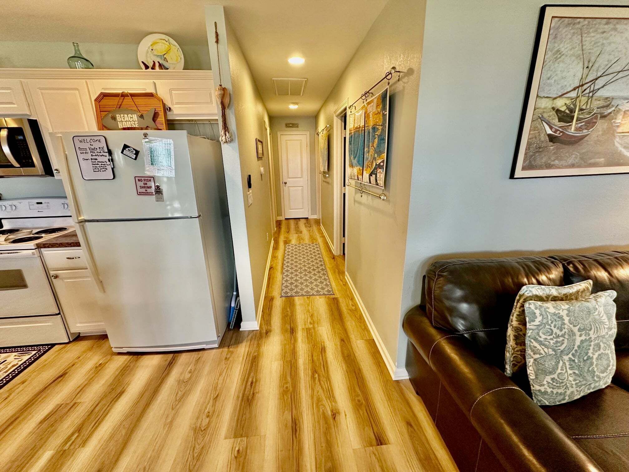 A narrow hallway with wooden flooring, a kitchen area on the left, and a brown leather sofa on the right.