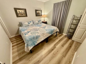 Une chambre à coucher avec un lit, deux tableaux encadrés au mur, une table de chevet avec une lampe, un rideau gris et une étagère en métal.