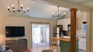 Ein modernes Wohnzimmer mit Flachbildfernseher, einer Couch, einem Couchtisch und einem Essbereich mit Holztisch und Stühlen. Es gibt eine Küchenzeile mit weißen Schränken und einer grünen Kücheninsel.