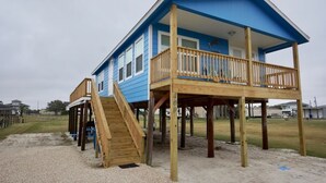 Une maison de plage à deux étages, dotée d’une terrasse en bois et d’un balcon, construite sur pilotis au-dessus d’un sol sablonneux.