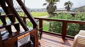 Een houten terras met een 'KEEP OUT' bord, een uitzicht op een parkeerplaats en palmbomen.