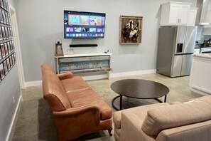 A modern living room with a brown leather sofa, a beige sofa, a small round table, a television mounted on the wall, and a refrigerator in the background.