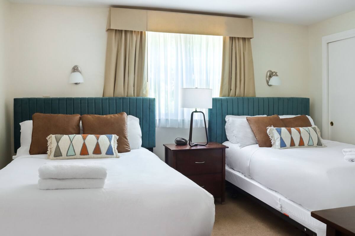 A hotel room with two beds, a nightstand, a lamp, and a window with curtains.