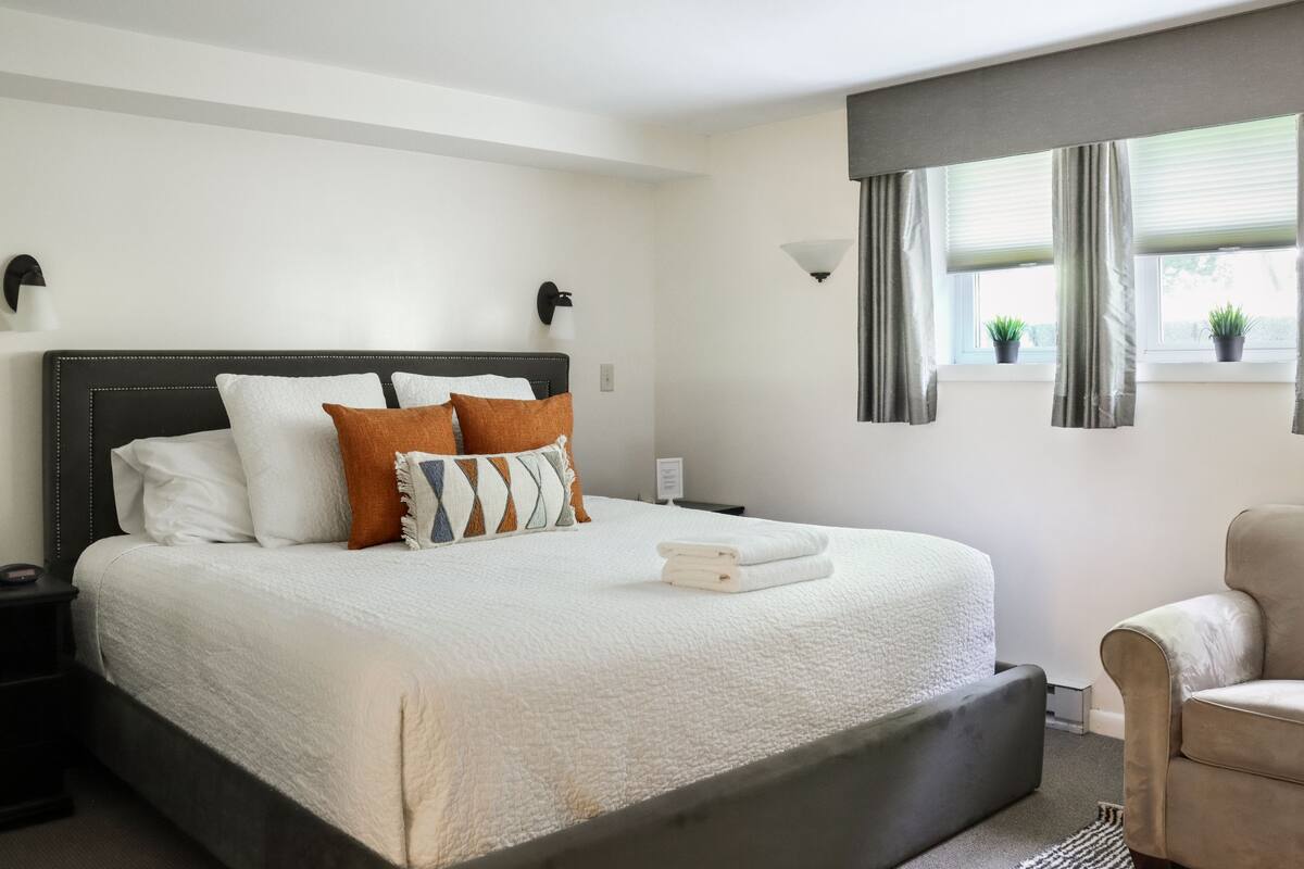 A neatly made bed with a white comforter and several pillows, a bedside table with a lamp, and a window with curtains and two potted plants.