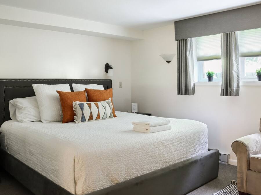 Ein ordentlich bezogenes Bett mit weißer Decke und mehreren Kissen, ein Nachttisch mit Lampe sowie ein Fenster mit Vorhängen und zwei Blumentöpfen.