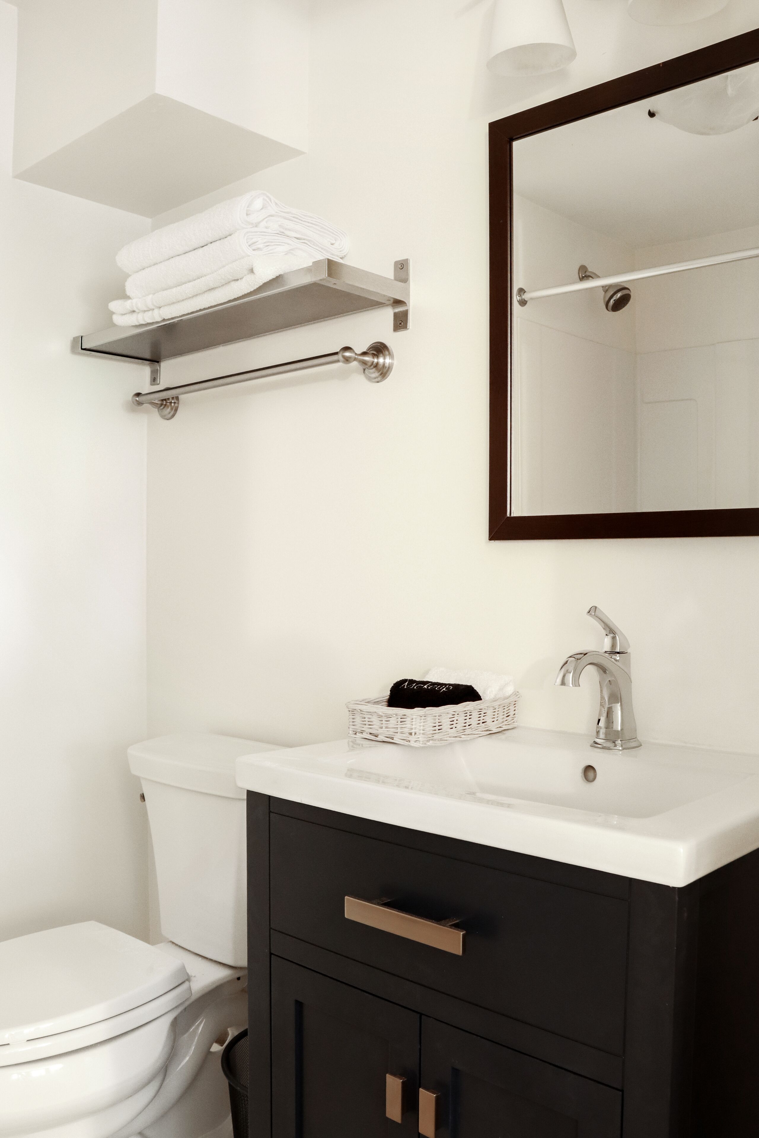 A modern bathroom with a white toilet, a black vanity with a silver faucet, a white sink, and a mirror above the sink. There is a towel rack above the toilet and a basket on the vanity.
