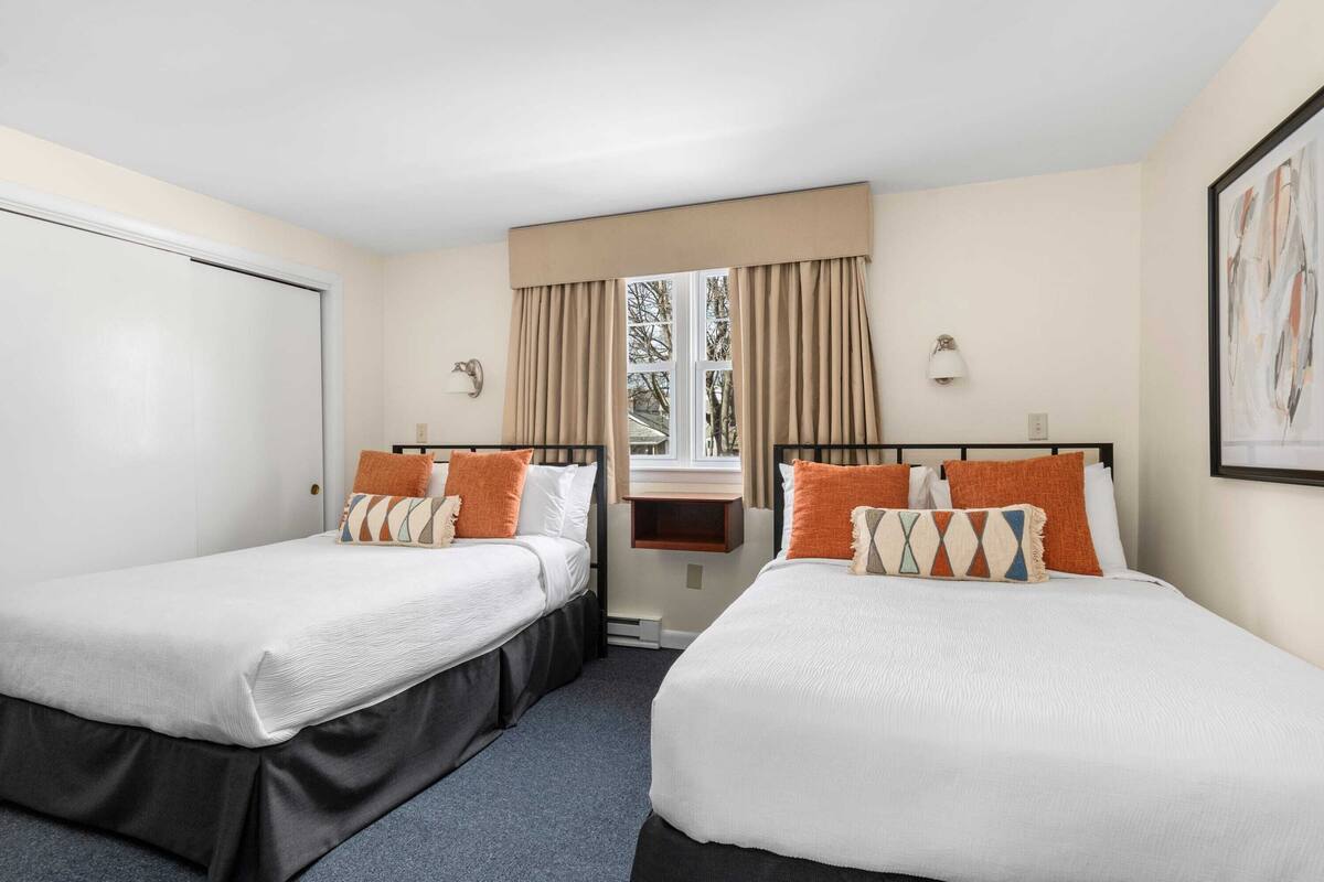 A hotel room with two single beds, a window with curtains, a wall-mounted lamp, and a framed picture.