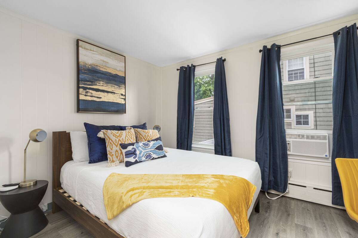 A bedroom with a bed, a bedside table, a lamp, a window with curtains, and a painting on the wall.
