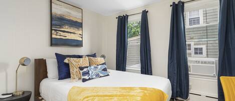 A bedroom with a bed, a bedside table, a lamp, a window with curtains, and a painting on the wall.