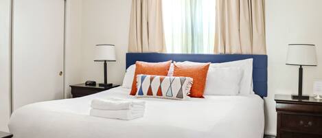 A neatly made bed with white linens and two pillows, one orange and one white, in a room with a large window and curtains.