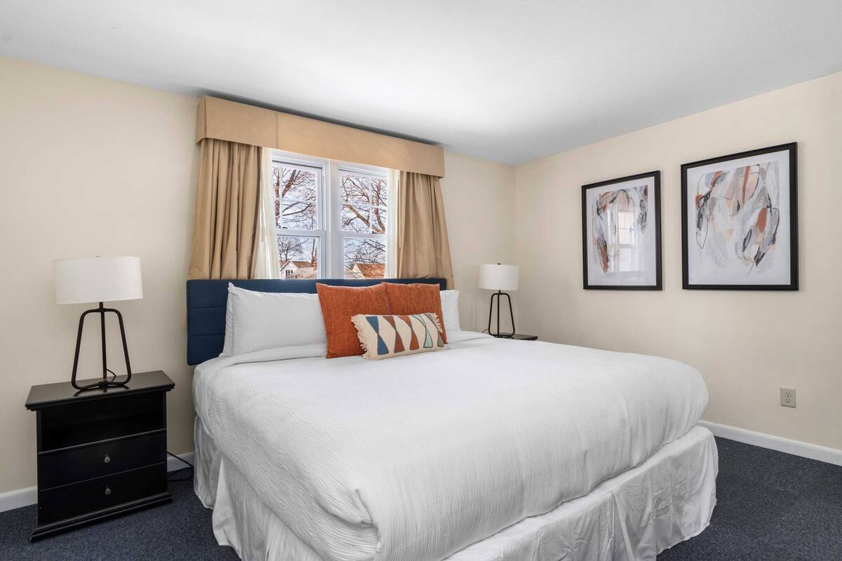 A neatly arranged hotel room with a large bed, two bedside tables, a window with curtains, and two framed abstract paintings on the wall.