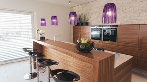 Una cocina moderna con una isla de madera, taburetes de bar, un frutero y lámparas colgantes moradas.
